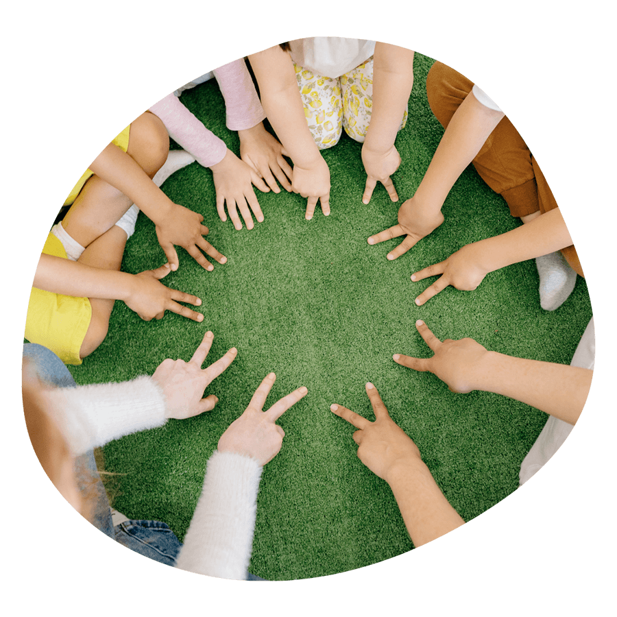 Un gruppo di bambini seduti in cerchio sull'erba con le mani al centro, durante un'attività CLIL del corso estivo Move Your Summer
