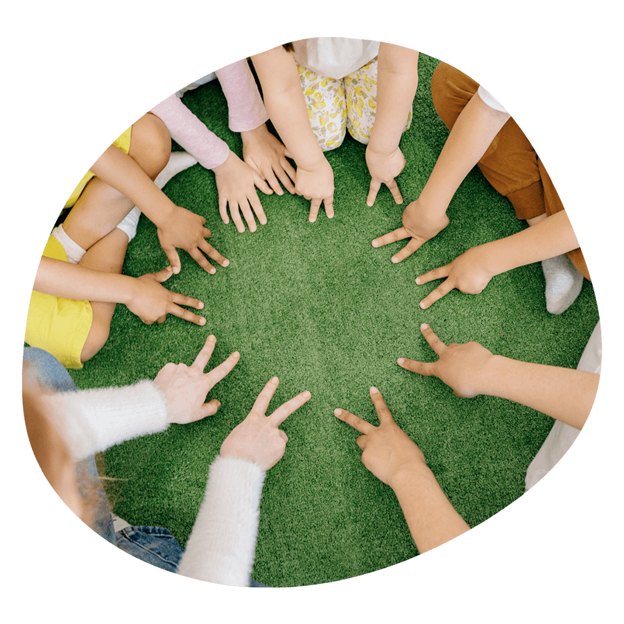 Un gruppo di bambini seduti in cerchio sull'erba con le mani al centro, durante un'attività CLIL del corso estivo Move Your Summer